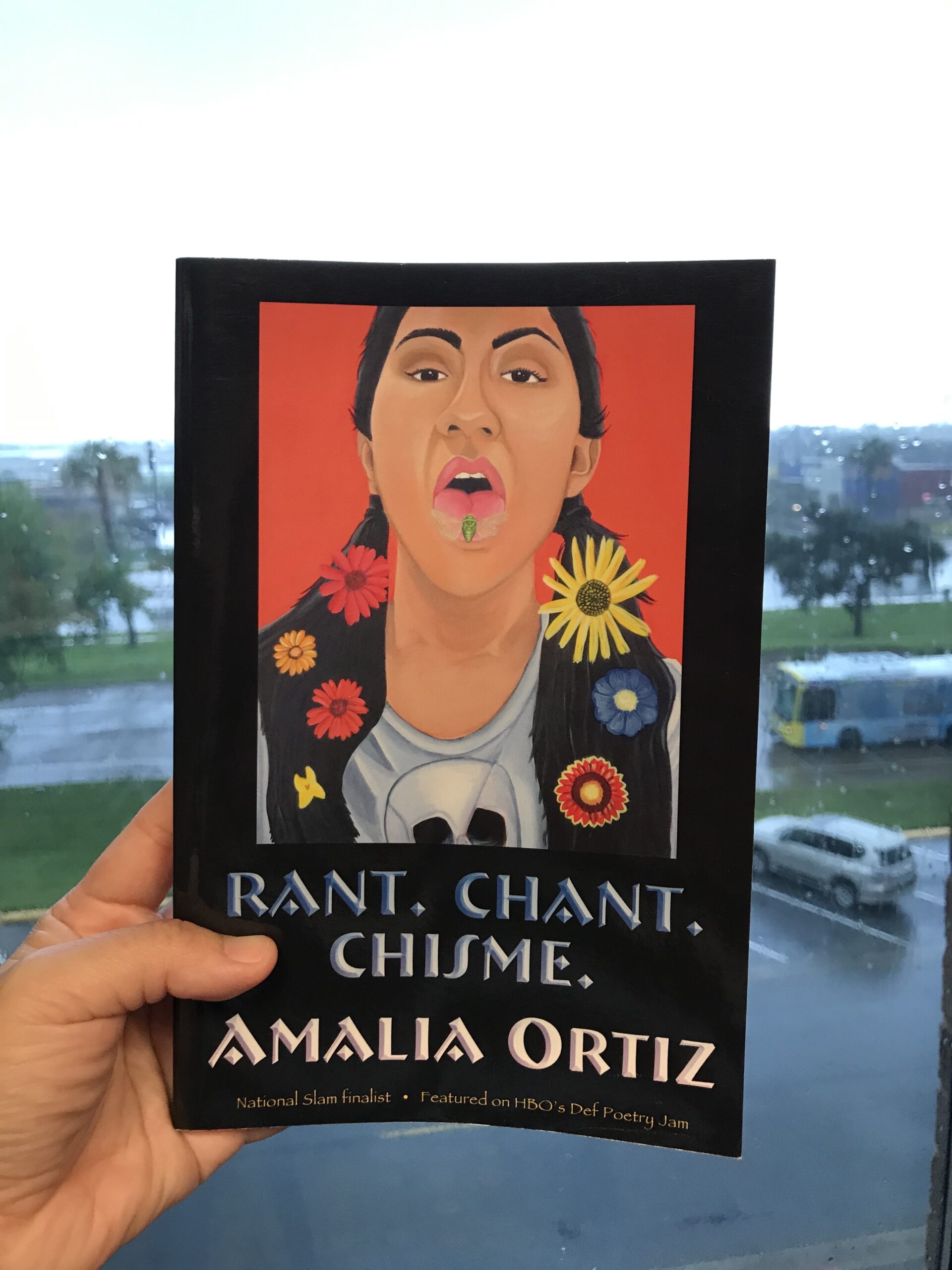 A hand holds up a copy of Amalia Ortiz's book, Rant. Chant. Chisme. The cover depicts a woman sticking her tongue out to reveal a cicada. Her dark hair is parted and features different brightly colored flowers. Source: Marlene Galván