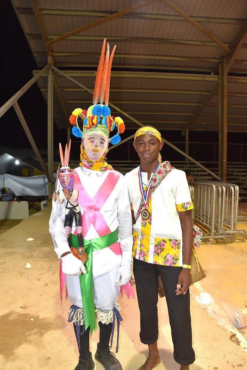 A Wanaragua dancer in white clothes, white mask and colorful accents stands next to a drummer wearing more normal clothes